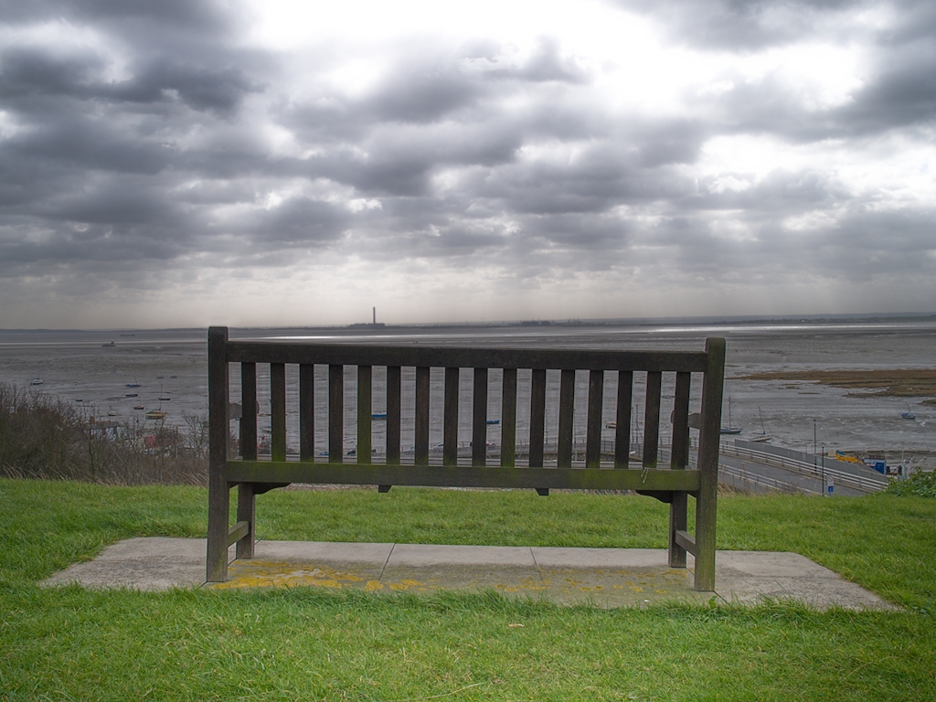 Wooden bench overlooking estuary with power station in far distance and dramatic sky.