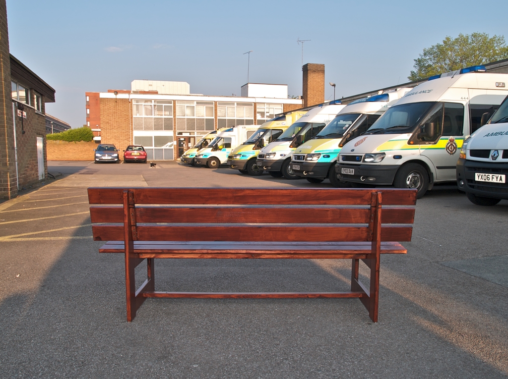 Wooden bench ovelooking ambulance station car park. Row of ambulances and office buildings in background.