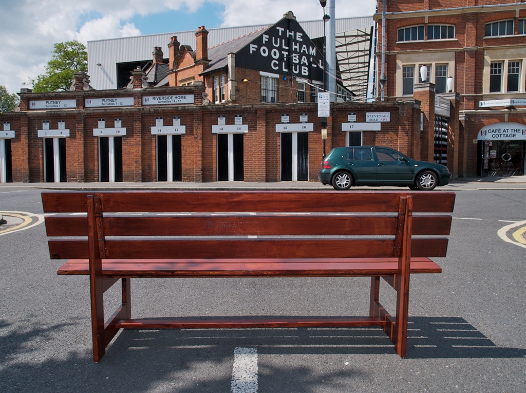 Wooden bench in road overlooking entrance to fulham football club with craven cottage in background.