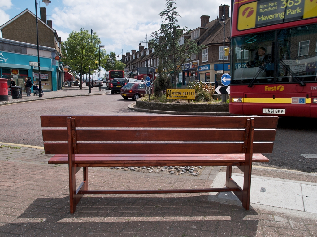 Wooden bench on edge of roundabout with red bus passing and row of shops in background.